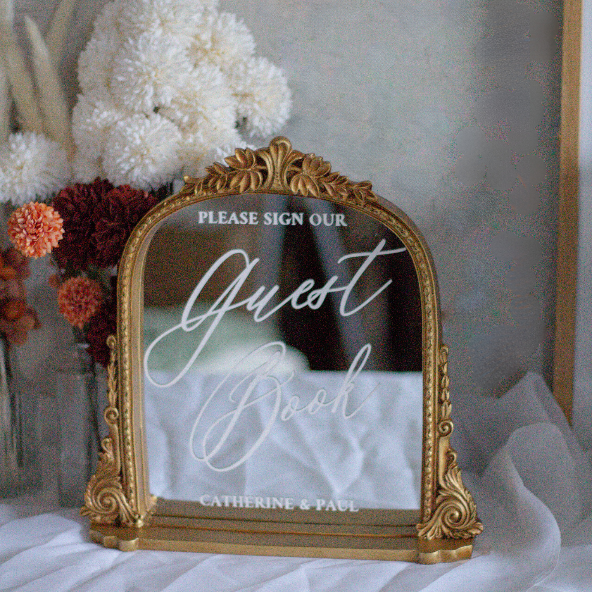 Elegant wedding guest book sign with gold frame and calligraphy font on a mirror, accented by floral decor.