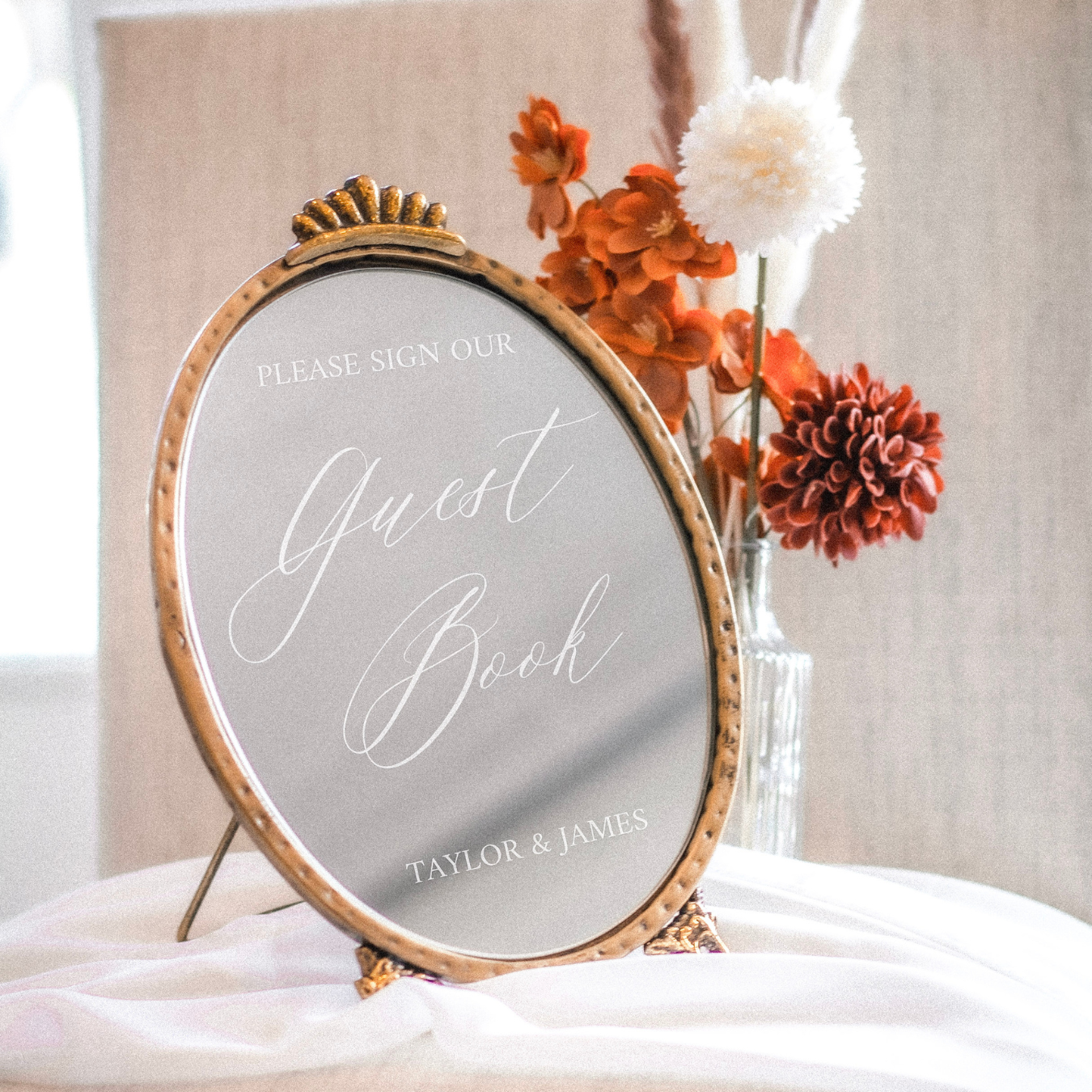Elegant wedding guest book sign on a decorative mirror with floral arrangement.
