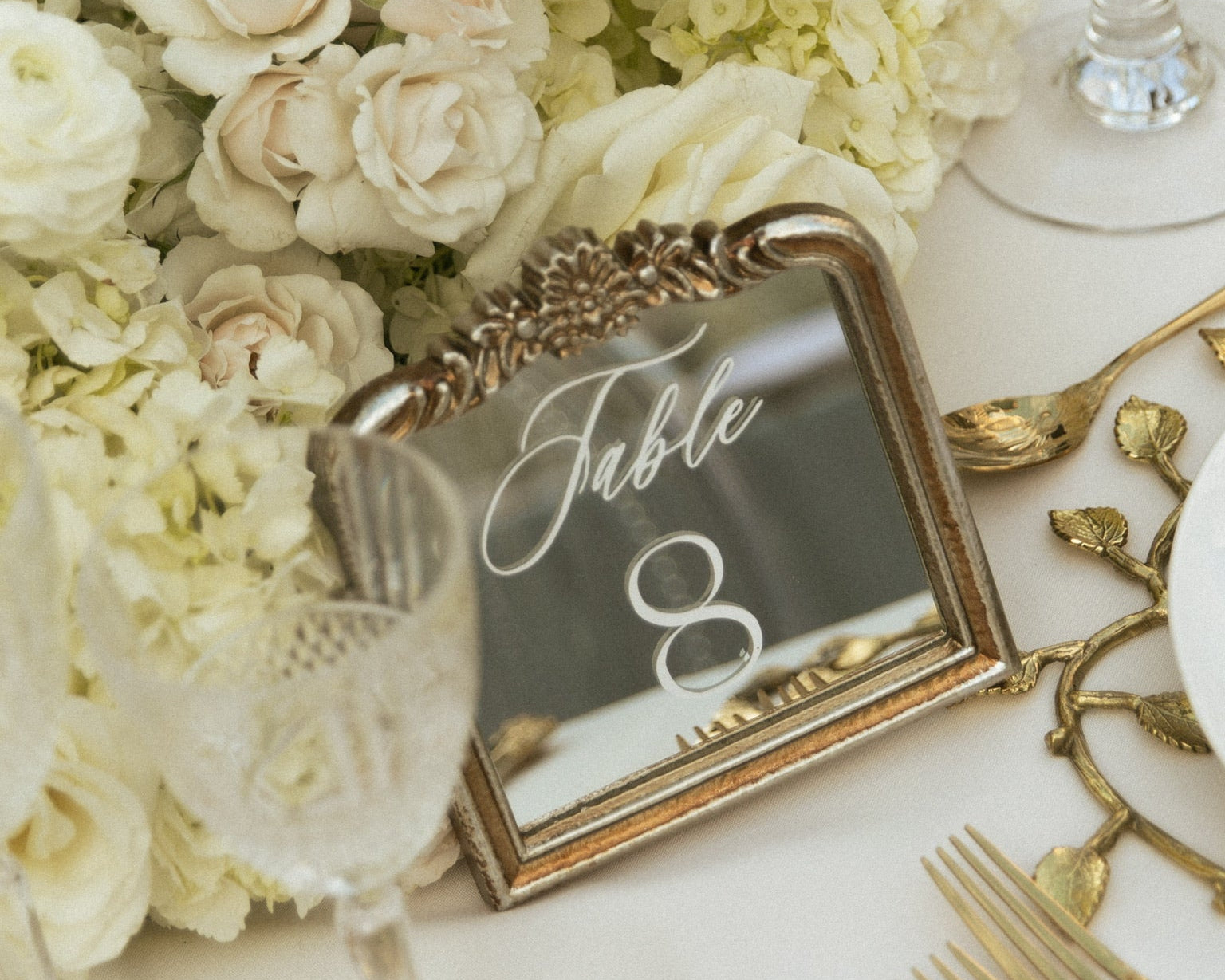 An ornate gold-framed table number sign reading 'Table 8' with a reflective mirror surface, styled on a white tablecloth alongside white flowers, clear glassware, and gold-toned flatware.