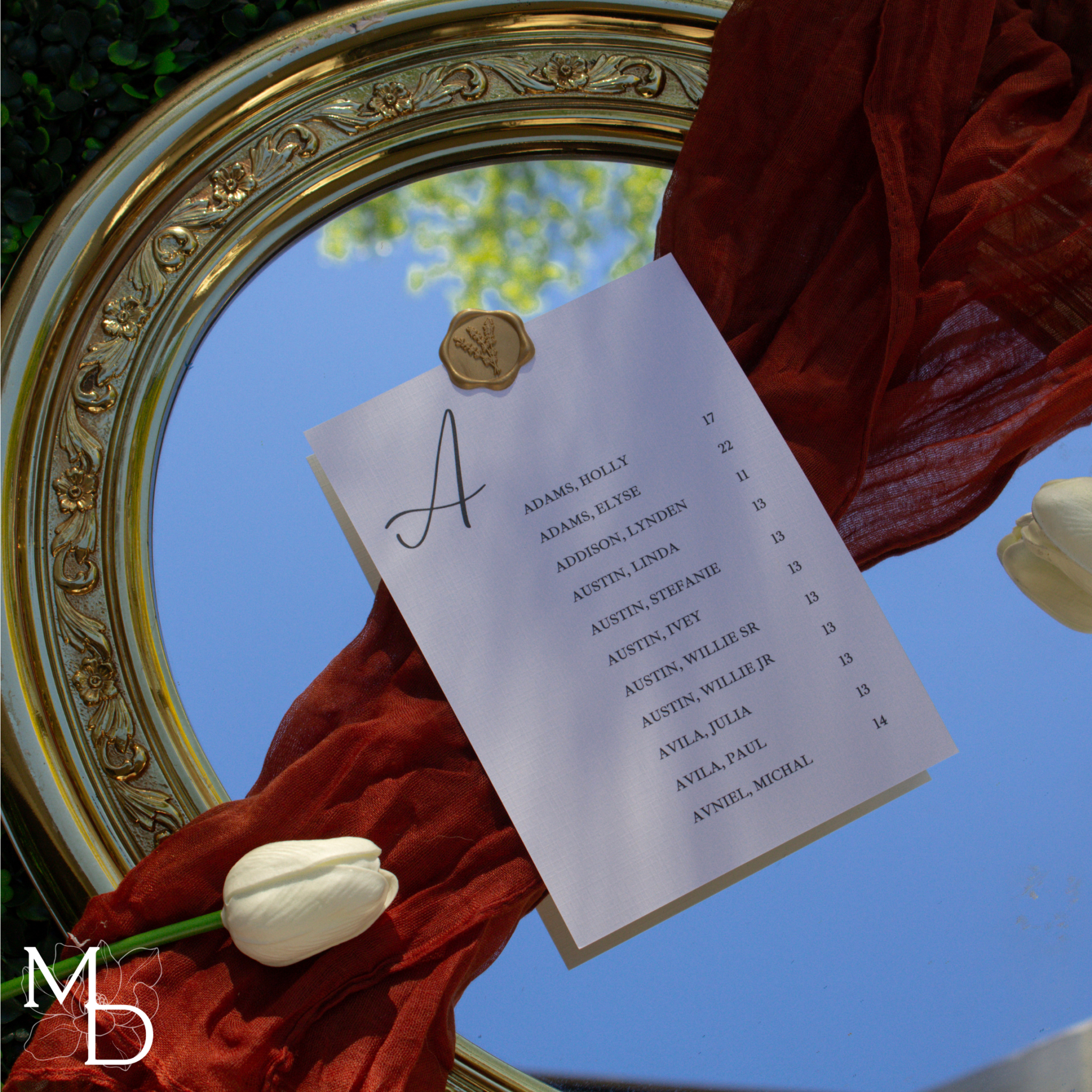 Elegant seating chart assignments card with wax seal, placed on a decorative mirror with a red cloth and white tulip accents.