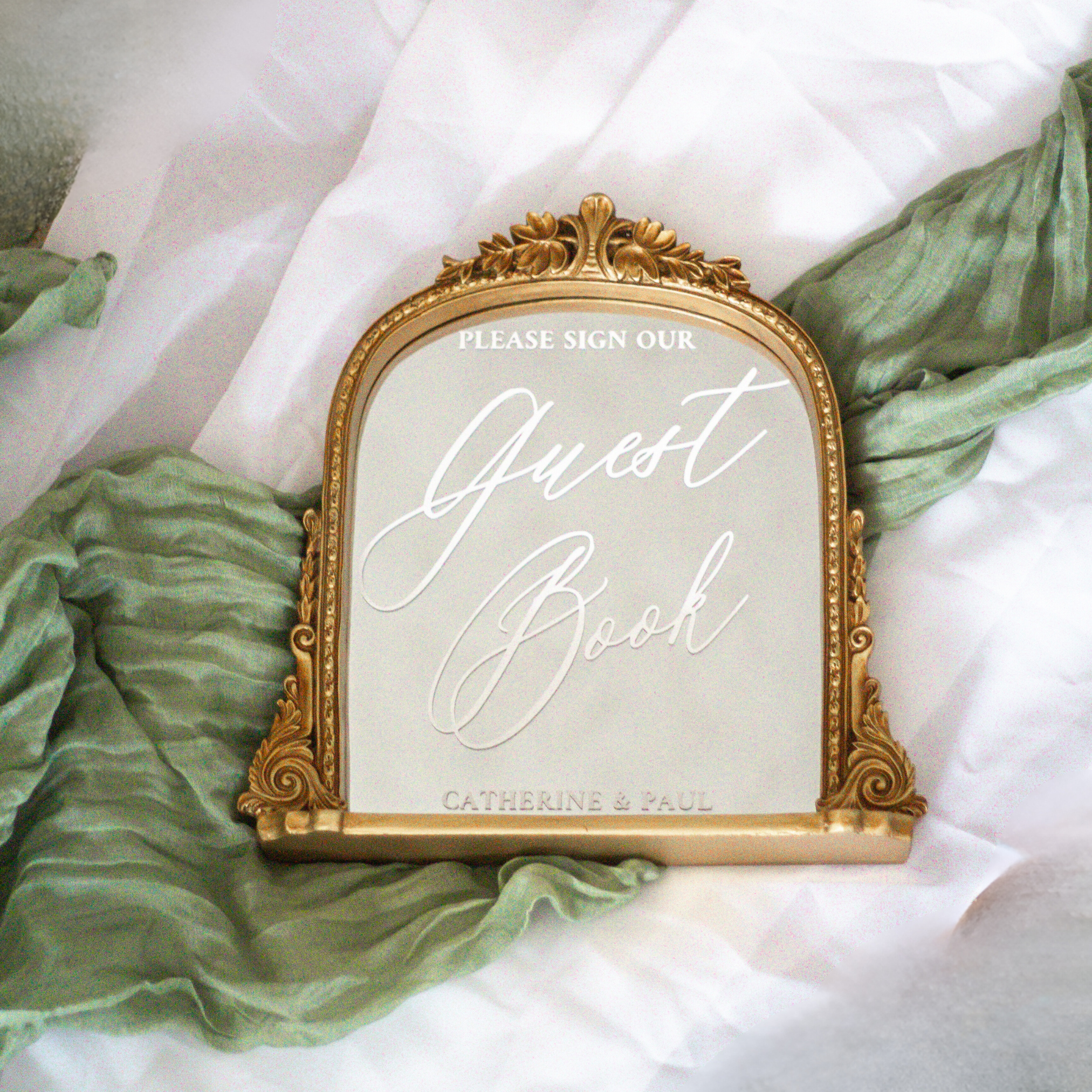 Elegant wedding guest book mirror sign with gold frame on white and green fabric.