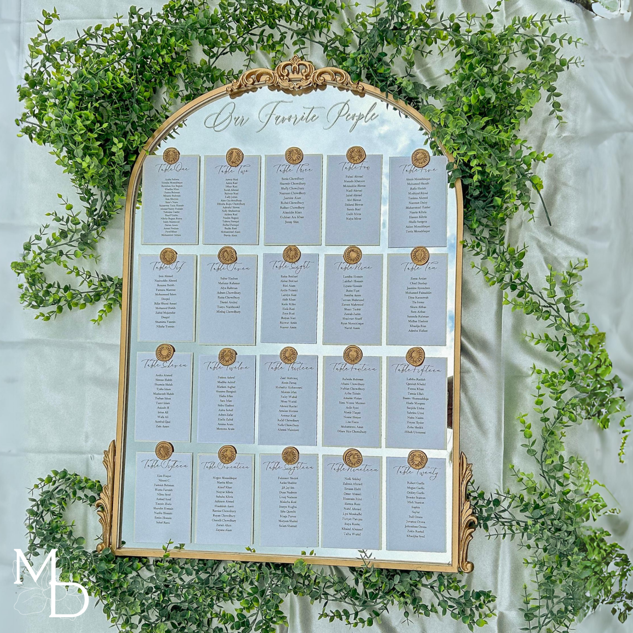 Elegant wedding guest table layout seating chart on gold arch mirror with greenery.