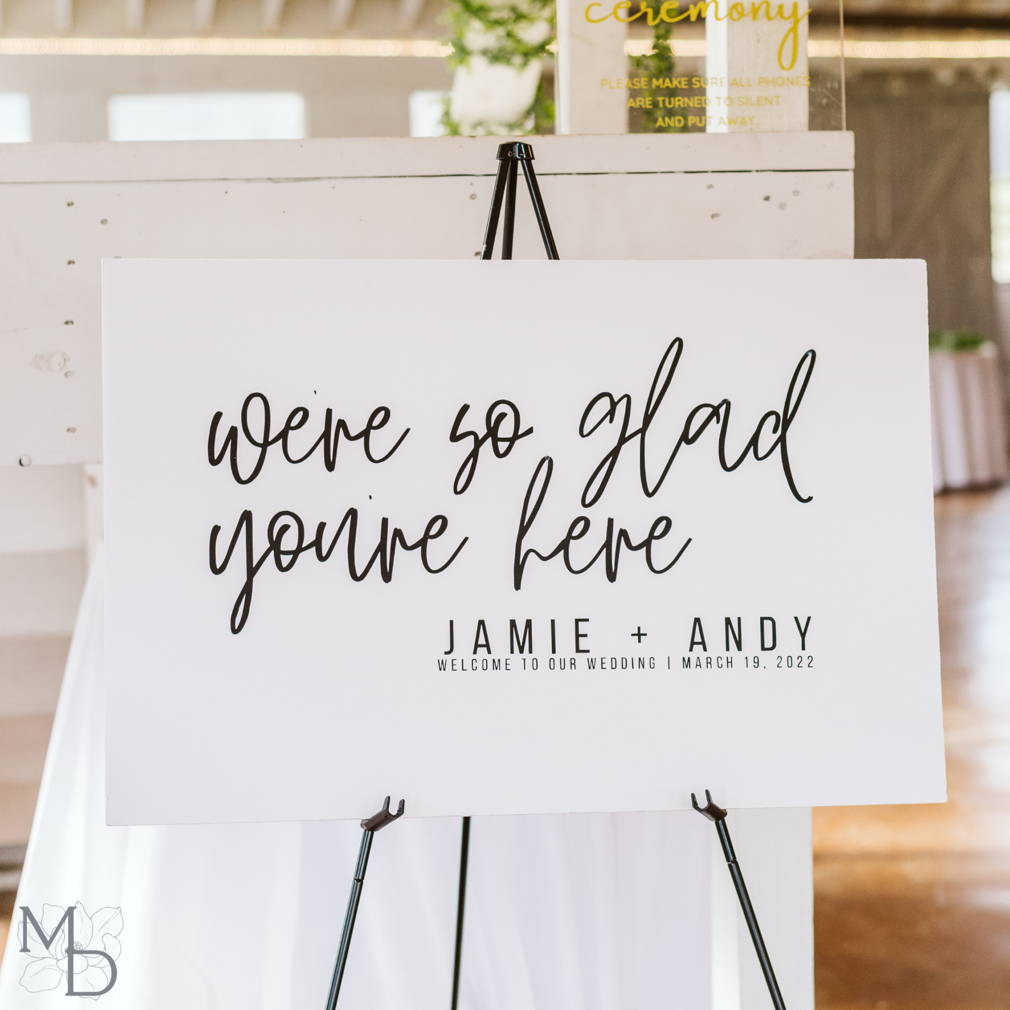 Minimalist wedding welcome sign with "We're So Glad You're Here" text on a foam board displayed on an easel.