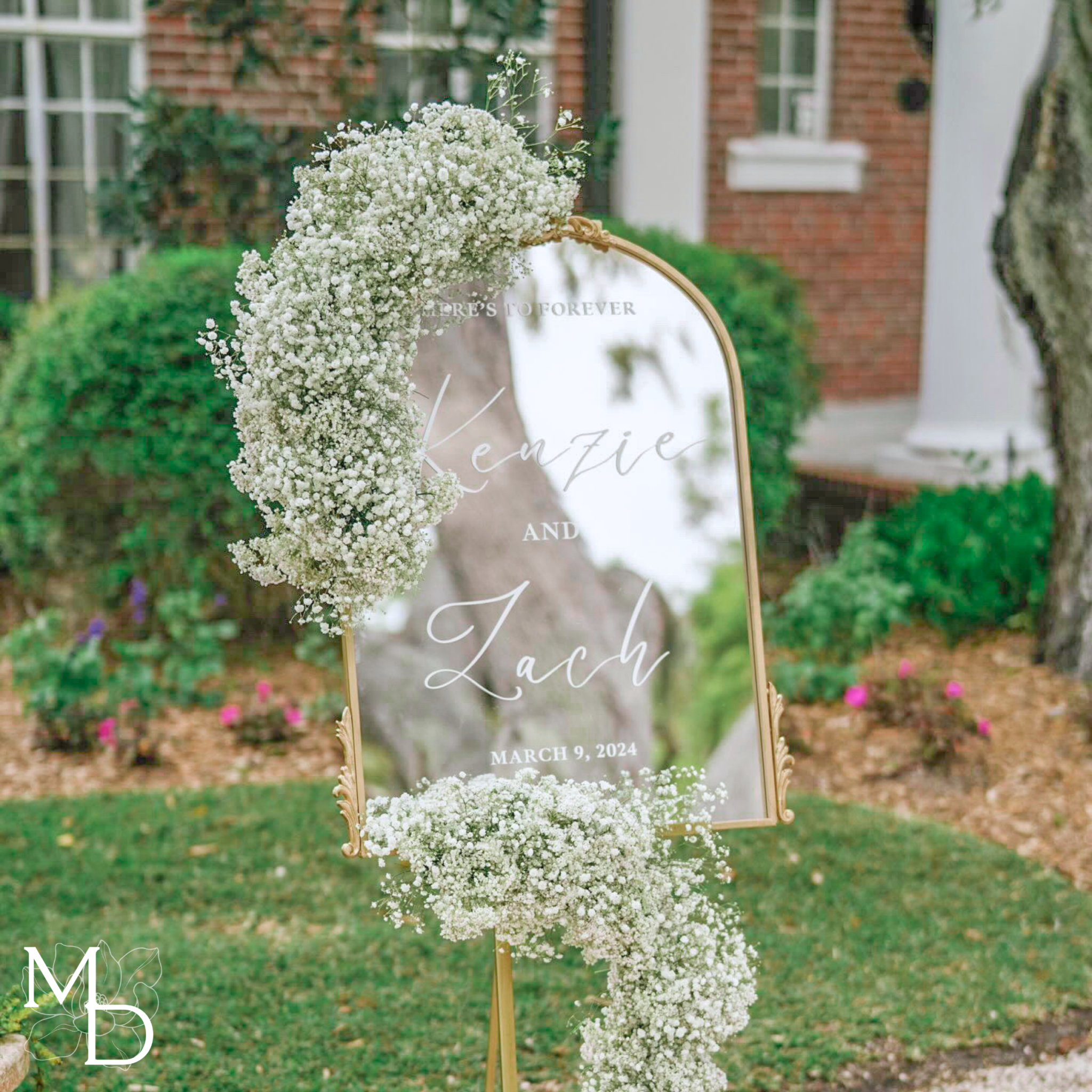 Elegant gold arch mirror wedding welcome sign with floral decoration in outdoor setting.