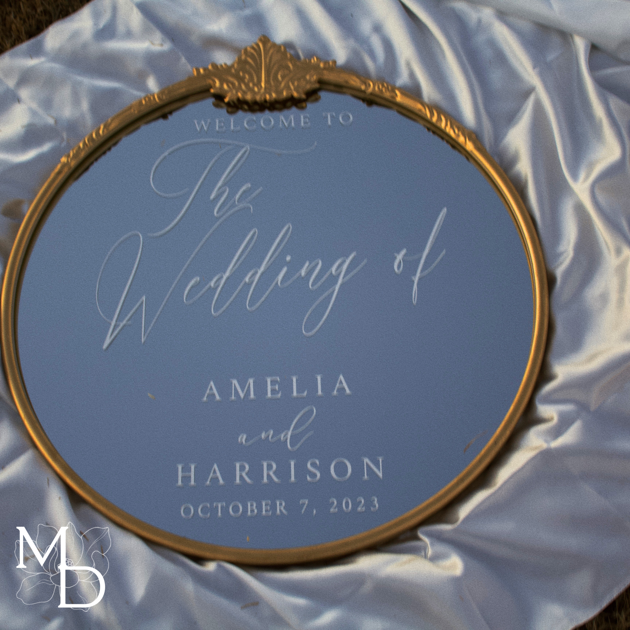 Gold ornate round mirror with custom wedding welcome sign on white cloth.