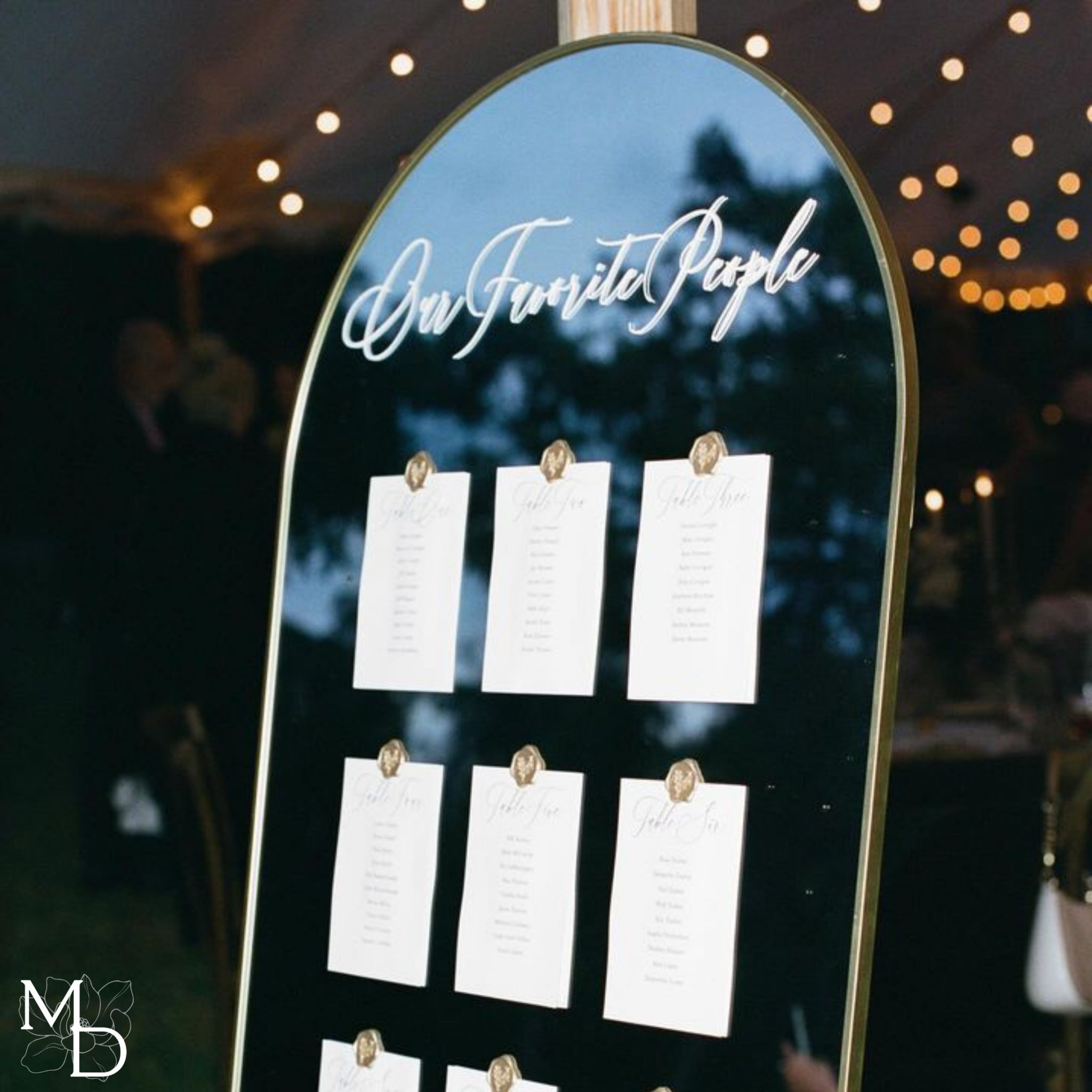 Large gold arch mirror with wedding table seating chart, displaying guest assignments on premium linen cardstock.