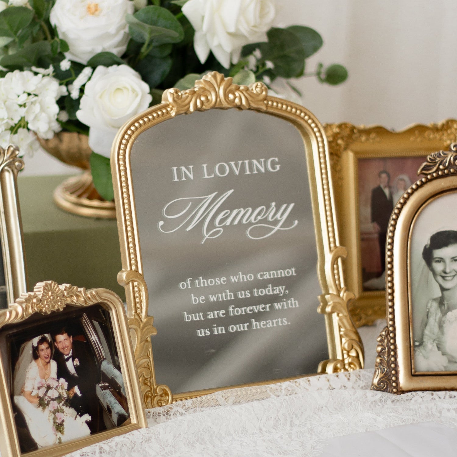 An ornate gold-framed memorial photo frame with the words 'IN LOVING Memory' displayed on a white lace-covered table, alongside other vintage-style picture frames and a white rose bouquet in the background.